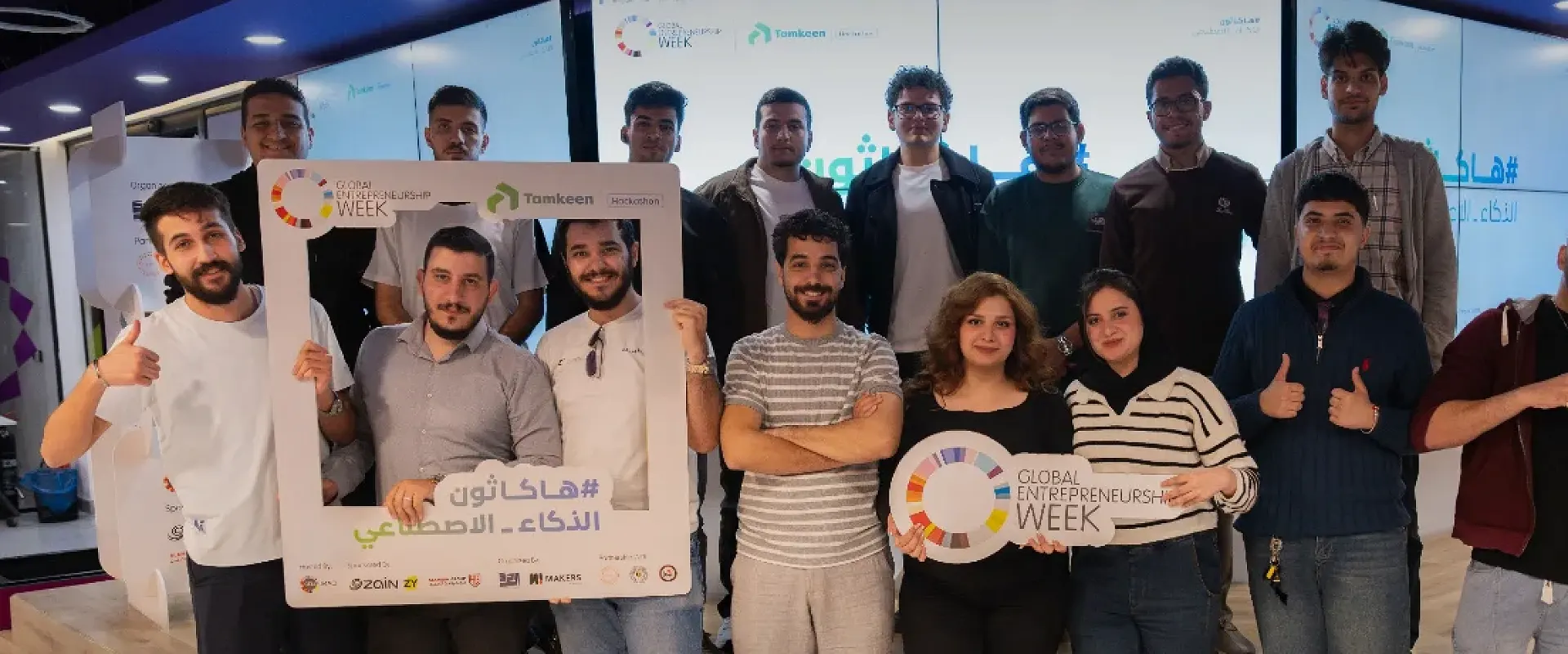 Participants in the AI Hackathon run by Makers of Baghdad during Global Entrepreneurship Week 2025 pose for a group photo, some holding event frames.